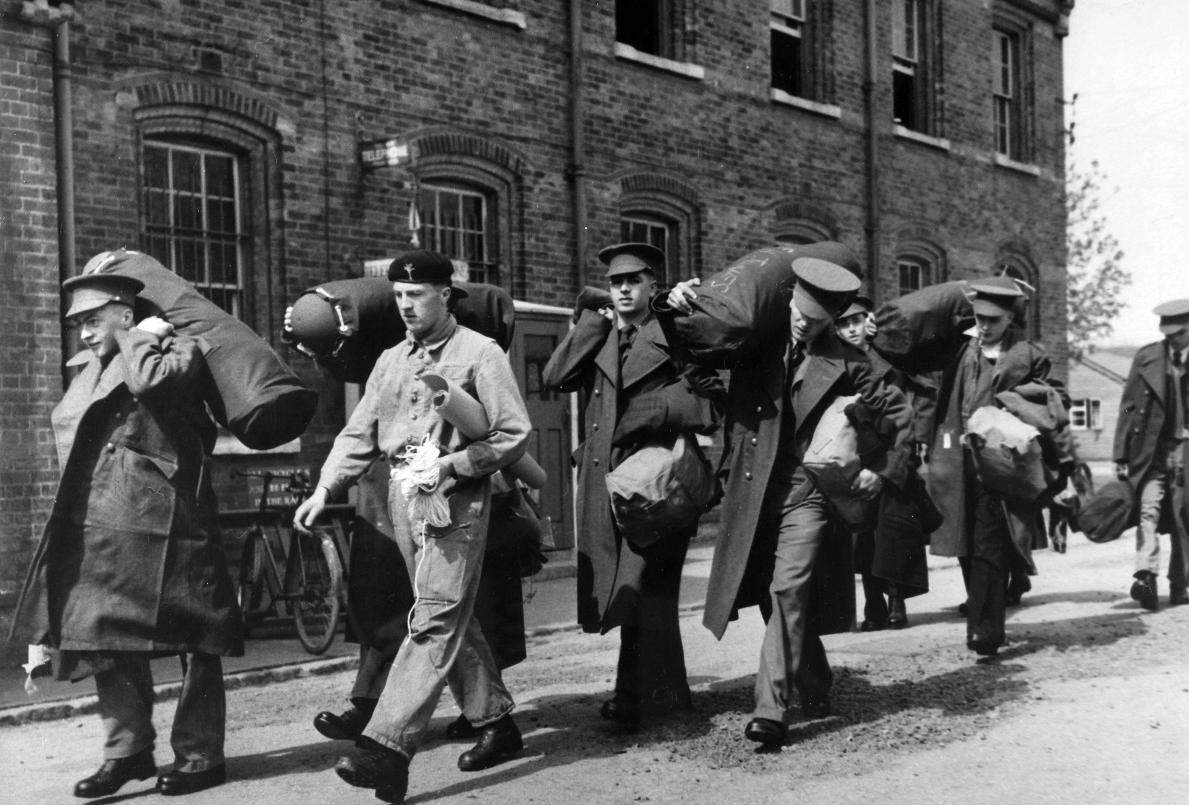 Black and white photo of National Service recruits walking in a line. They are carrying their belongings over their shoulders and in their arms. They are dressed in uniforms, with some wearing coats. There is a brick building behind them.