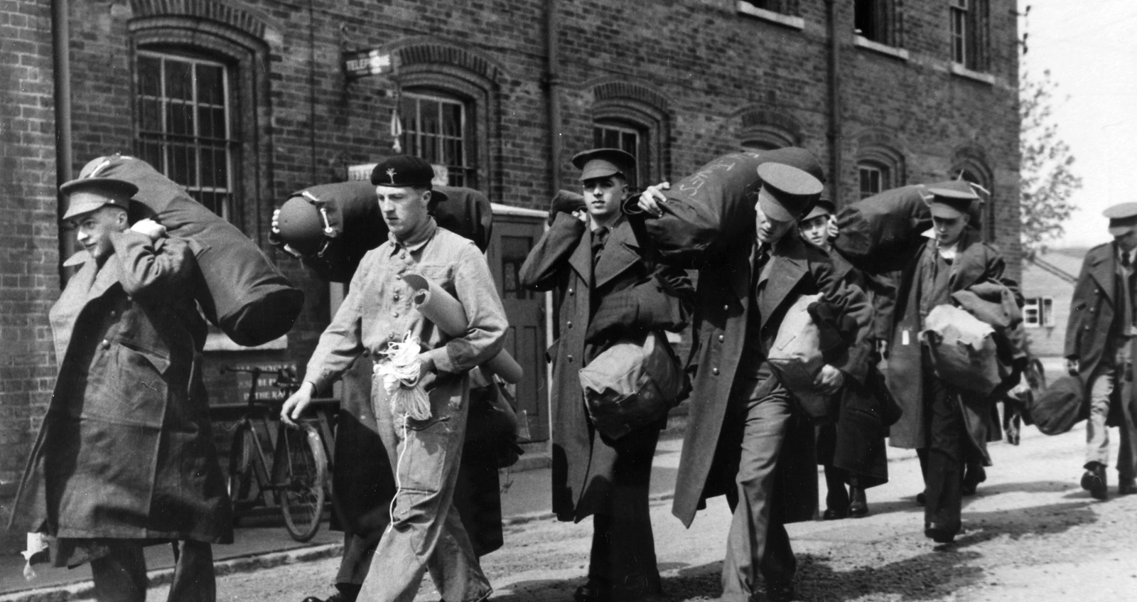 49472 Black and white photo of National Service recruits walking in a line. They are carrying their belongings over their shoulders and in their arms. They are dressed in uniforms, with some wearing coats. There is a brick building behind them.