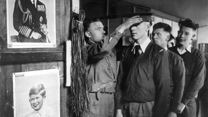 Black and white photo of a National Service recruit having his hat fitted. Others are lined up behind him. We can see some posters on the wall to the side of them.