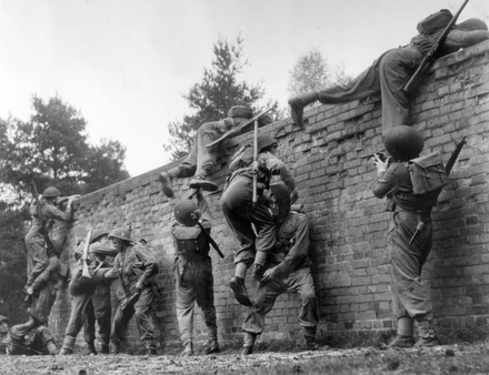 Black and white photo of National Service recruits scaling a nine foot brick wall. Some are being hoisted over the wall by others.