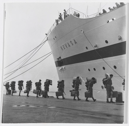 49477 Black and white photo of troops walking in a line about to board the troopship 'Nevasa'. The are carrying suitcases and other beloongings. We can see the ship behind them, with some people looking down from the top of the ship..