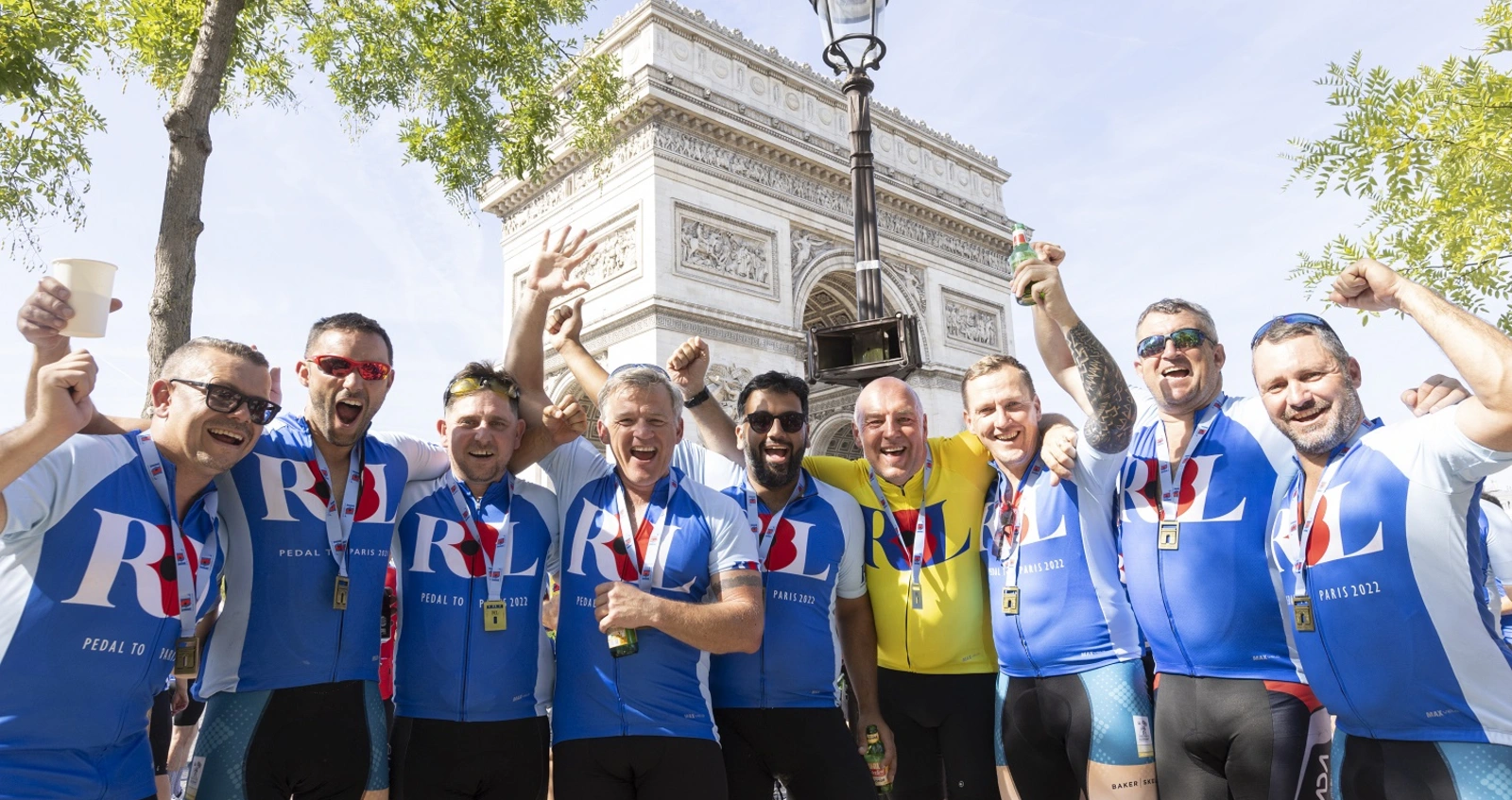 Pedal_to_Paris_2022_DAY4-125 A group of cyclists wearing blue RBL cycling jerseys celebrate arriving in Paris. They are stood in front of the Arc de Triomphe and raising their hands to cheer