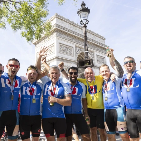 A group of cyclists wearing blue RBL cycling jerseys celebrate arriving in Paris. They are stood in front of the Arc de Triomphe and raising their hands to cheer
