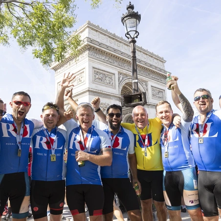 Pedal_to_Paris_2022_DAY4-125 A group of cyclists wearing blue RBL cycling jerseys celebrate arriving in Paris. They are stood in front of the Arc de Triomphe and raising their hands to cheer